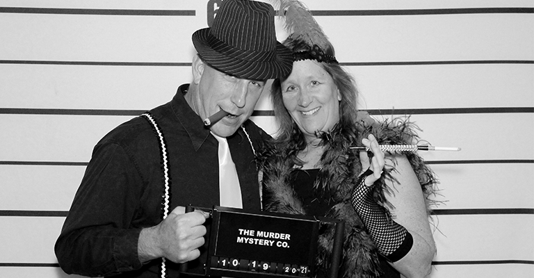 A black and white photo of a couple in a simulated mug shot holding a placard that reads 'The Murder Mystery Co.'