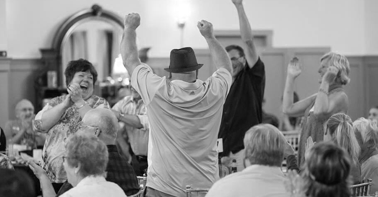 A black and white photo of people sitting in a dining room.  A person is standing with their back to the camera and has their arms up in celebration and several others are also standing and clapping